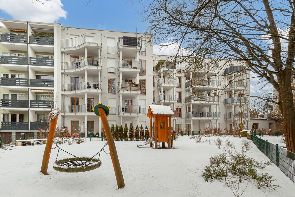 Schneebedeckter Spielplatz mit Klettergerüst und Schaukel vor Wohngebäuden.