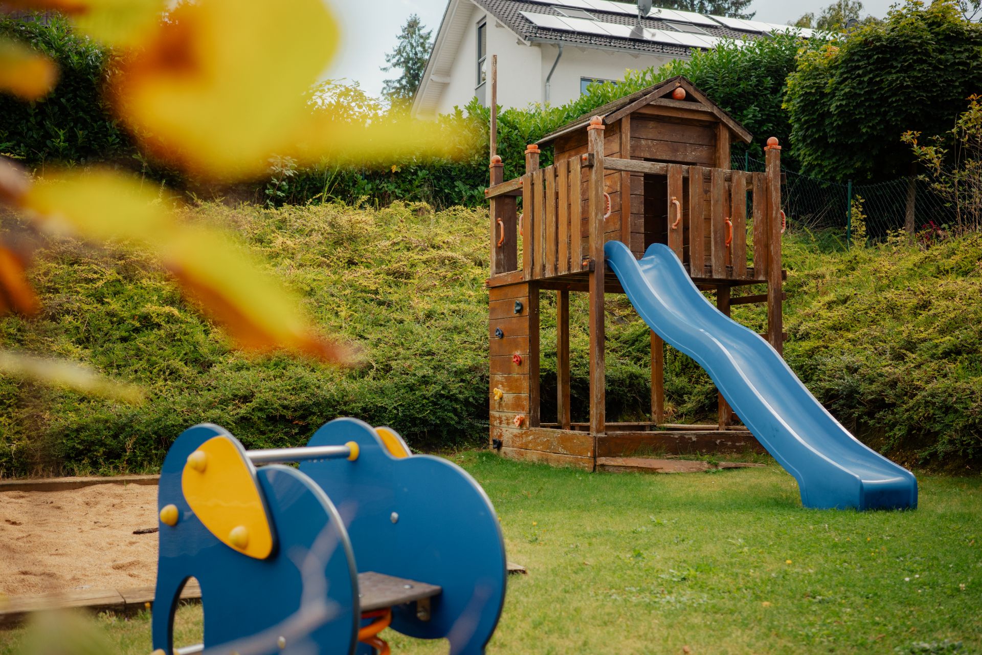Garten mit Holzspielhaus, blauer Rutsche und Wippe auf grünem Rasen.