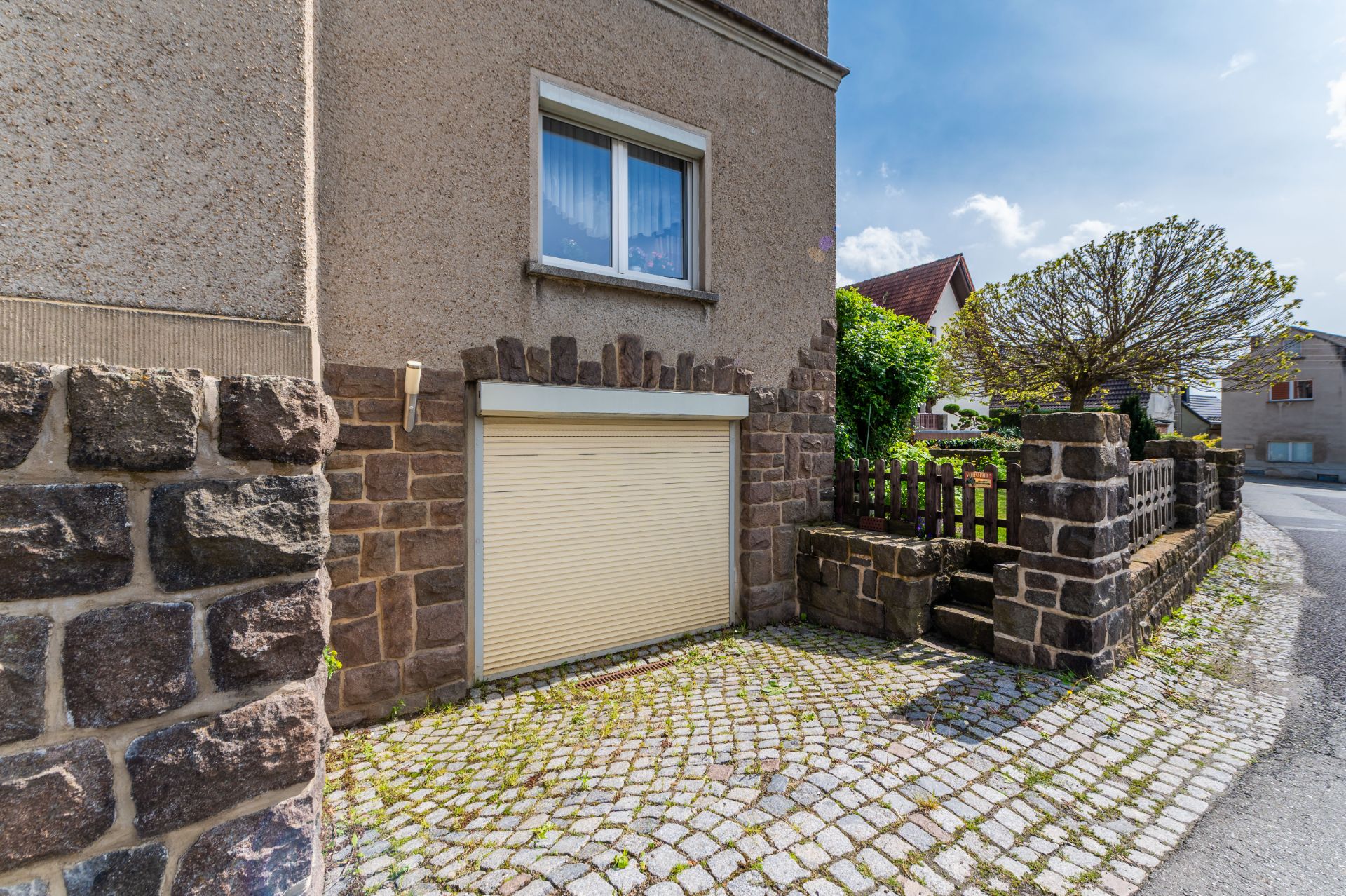 Garagentor mit gelbem Rollladen an einem Wohnhaus, umgeben von Steinmauer und Pflastersteinen.