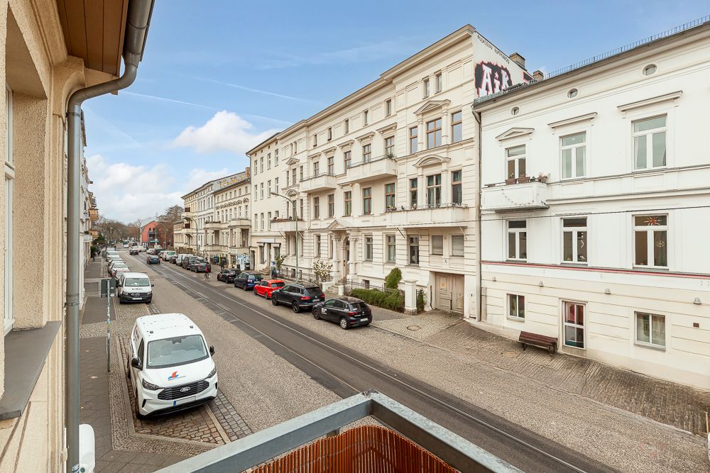 Blick von einem Balkon auf eine ruhige Straße mit geparkten Autos und mehrstöckigen Wohnhäusern.
