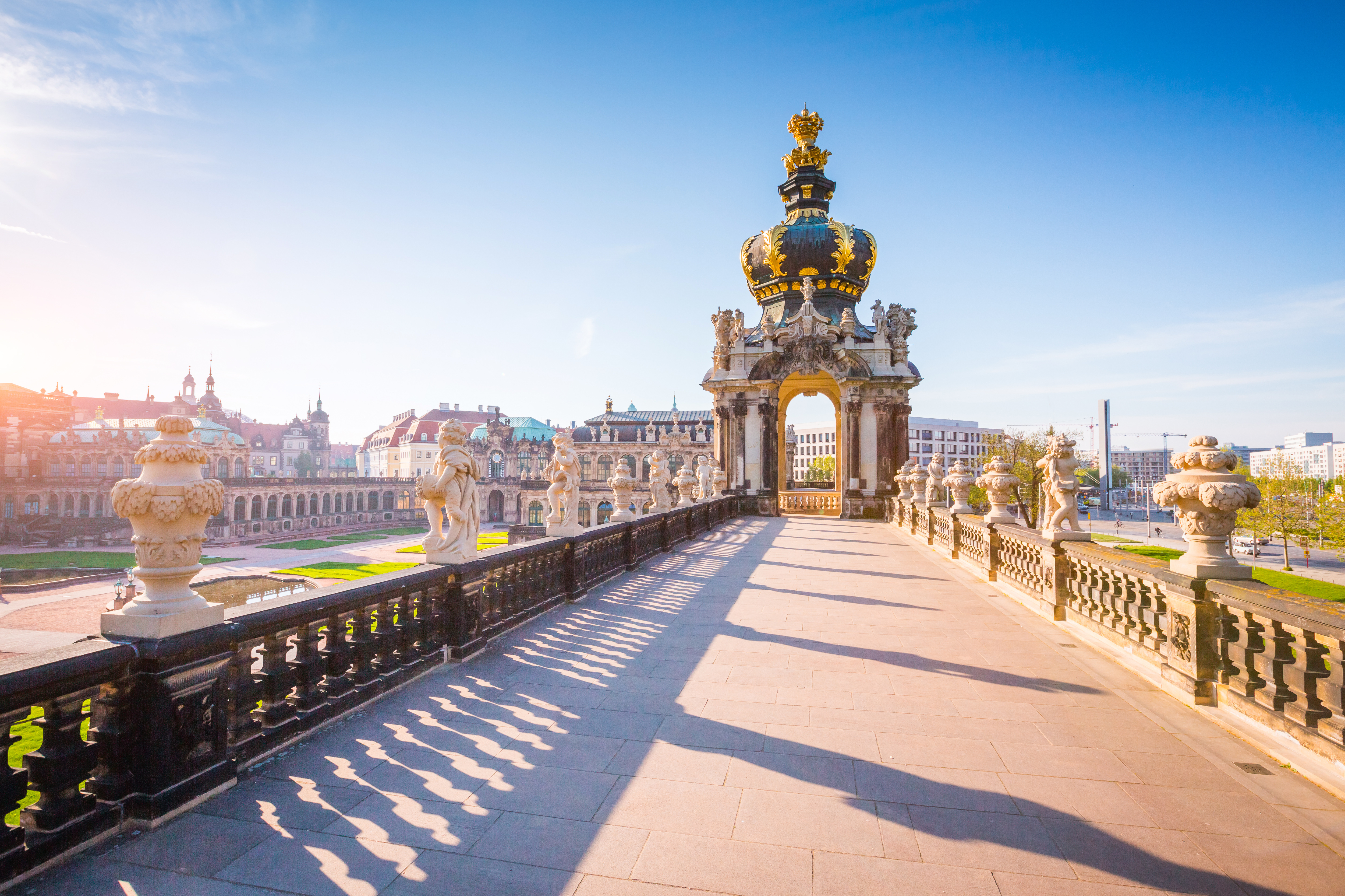 Steinerner Weg mit Balustrade und Skulpturen, im Hintergrund ein großes Tor mit goldener Krone.