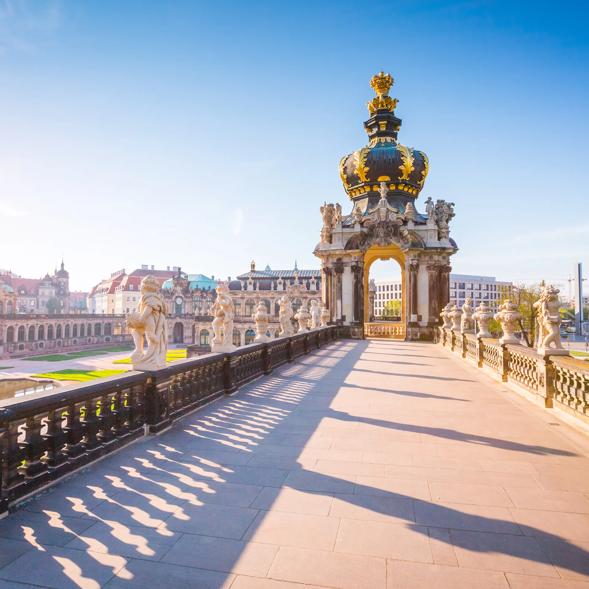 Steinerner Weg mit Balustrade und Skulpturen, im Hintergrund ein großes Tor mit goldener Krone.