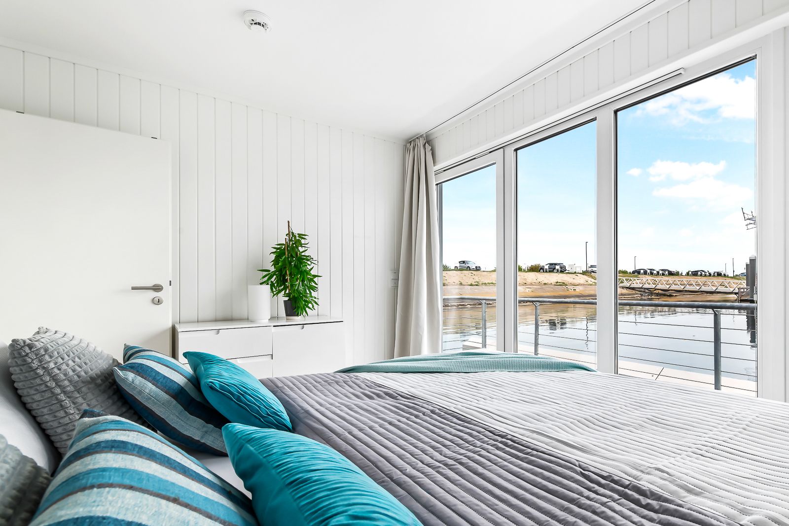 Helles Schlafzimmer mit großem Fenster, Bett, grauer Decke und blauen Kissen, Blick nach draußen auf Wasser.