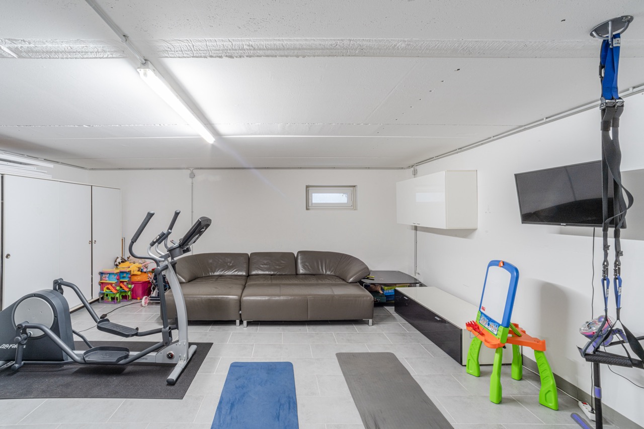 Heller Raum mit Sofa, Heimtrainer, Kinderspielzeug, Fernseher und Fitnessmatten.