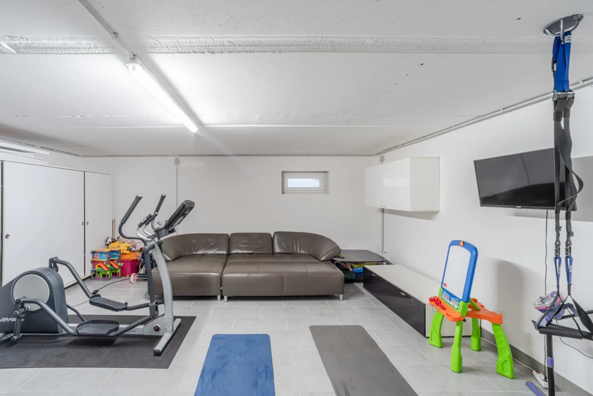 Heller Raum mit Sofa, Heimtrainer, Kinderspielzeug, Fernseher und Fitnessmatten.