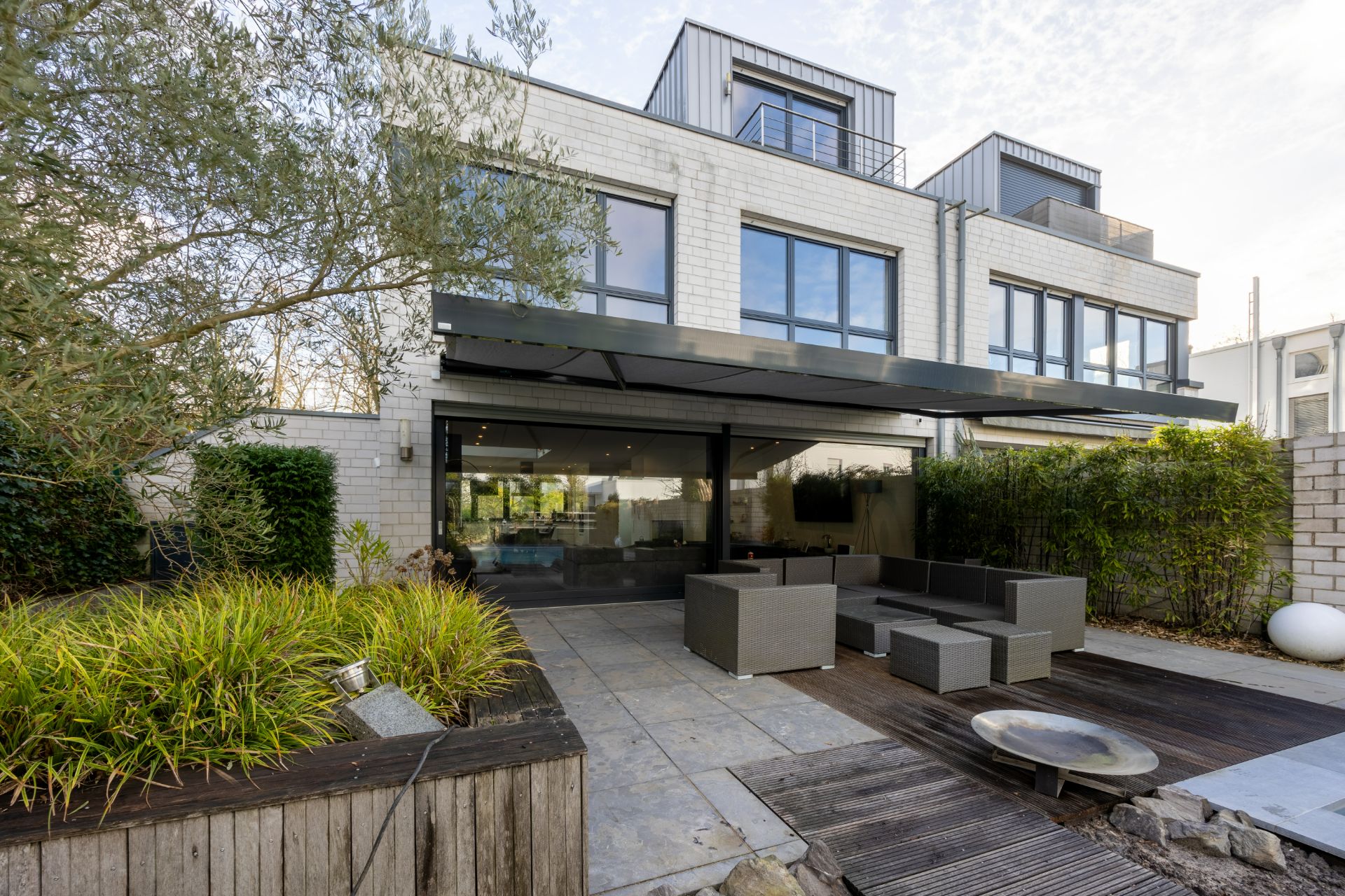 Moderne Terrasse mit Sitzmöbeln, Holz- und Steinboden, großer Fensterfront und begrünten Beeten.