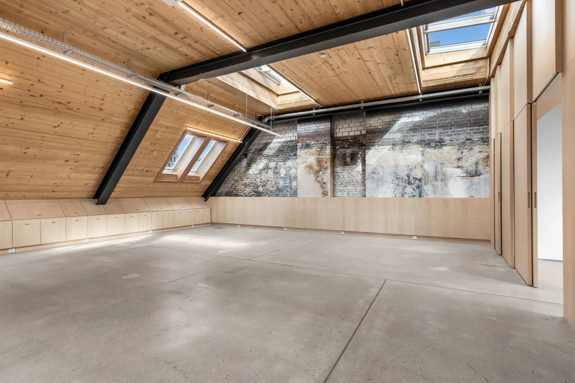 Heller Dachraum mit Holzdecke, Sichtbalken, Dachfenstern und offenliegender Backsteinwand.