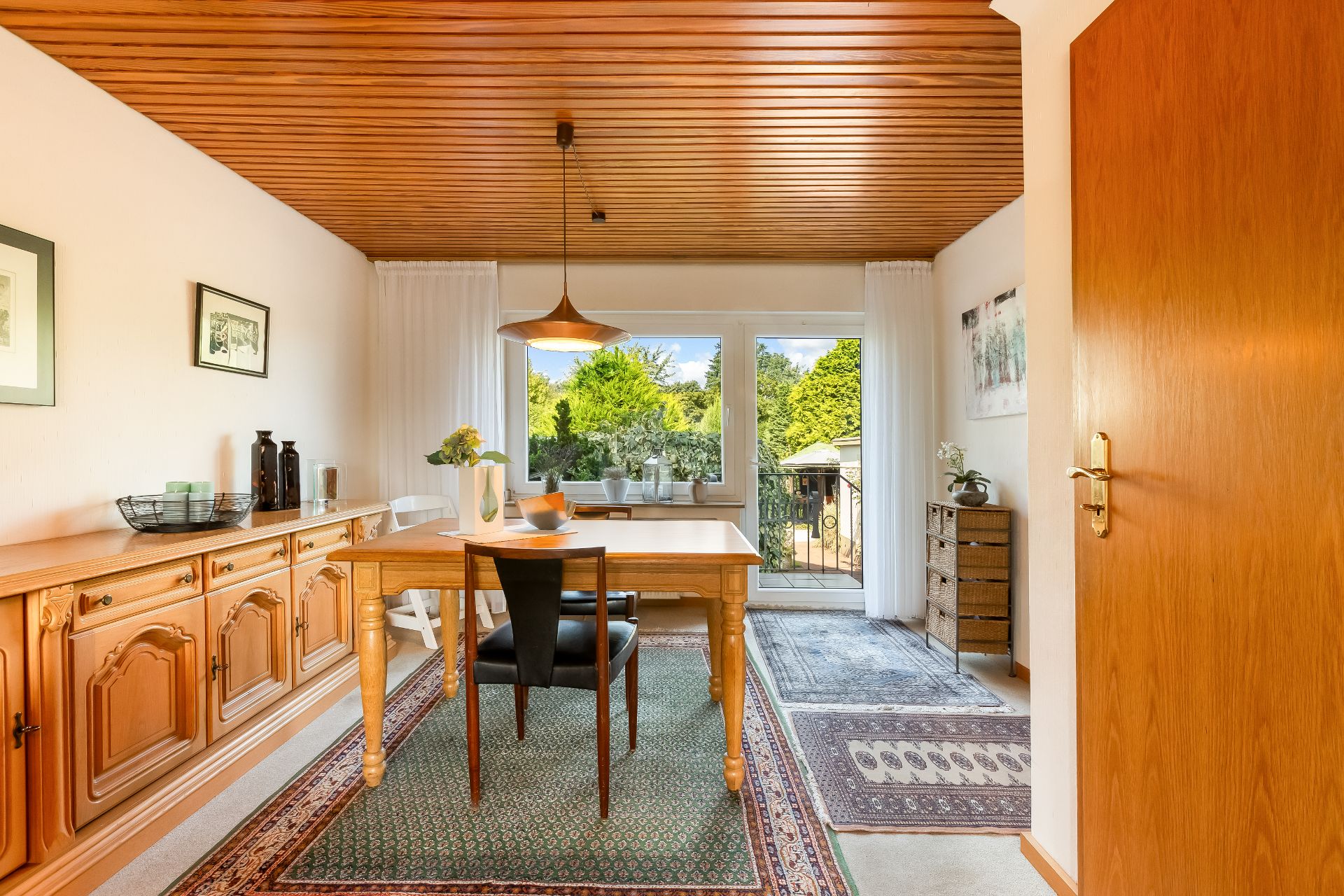 Esszimmer mit Holztisch, Teppichen und großen Fenstern mit Blick ins Grüne.