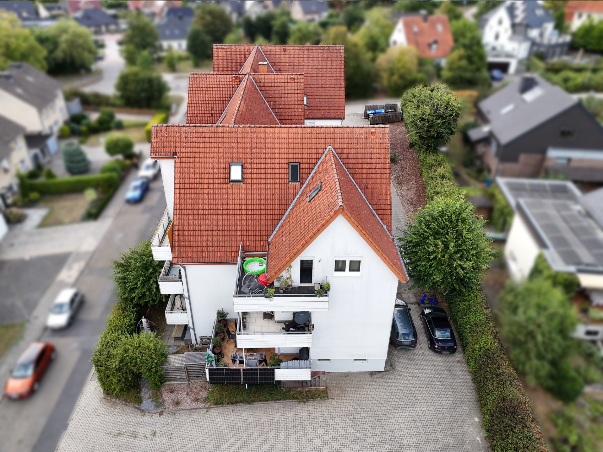 Luftaufnahme eines weißen Mehrfamilienhauses mit rotem Dach und Balkonen, umgeben von Bäumen.