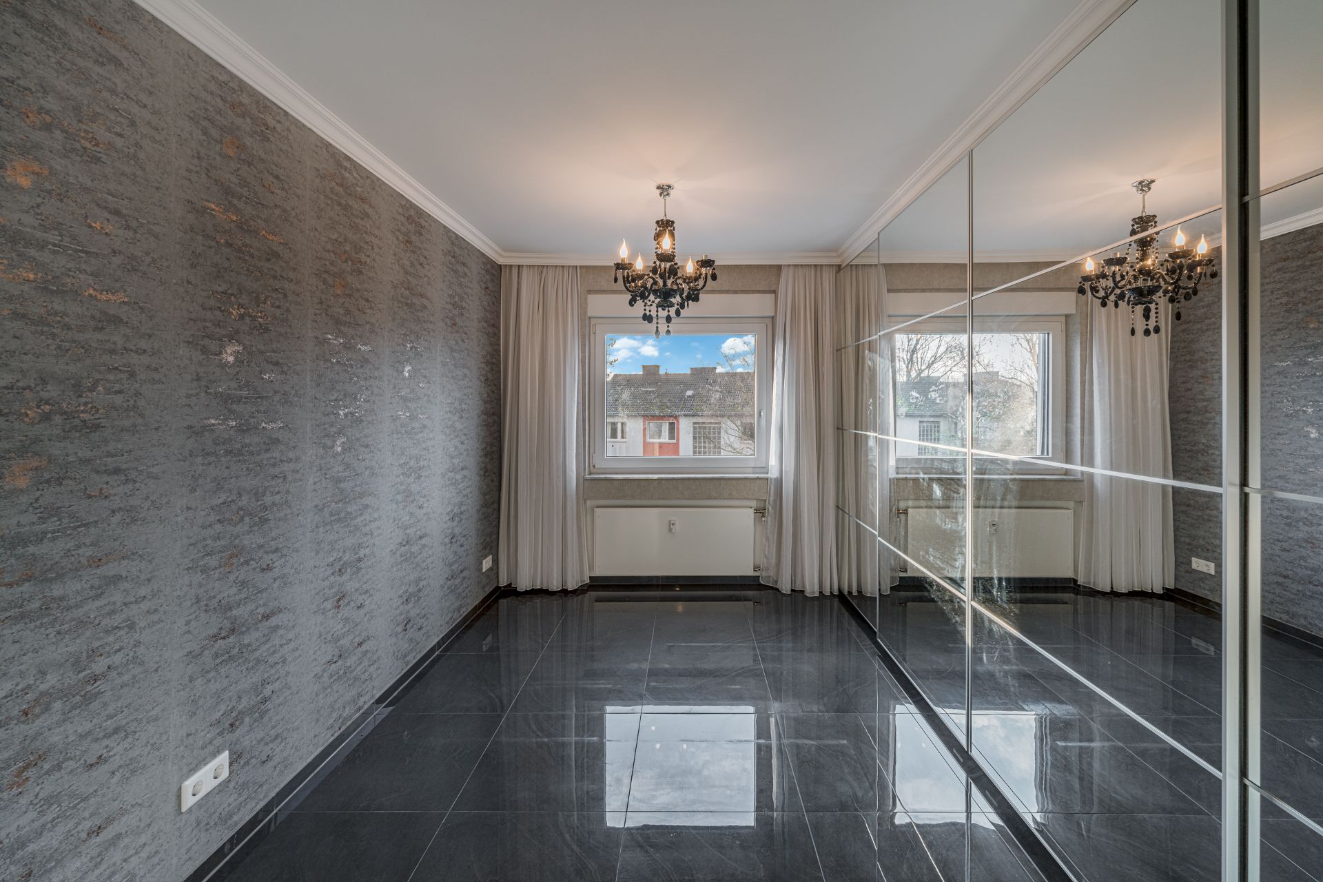 Leerer Raum mit grauem Fliesenboden, großem Spiegel, Fenster und Kronleuchter.