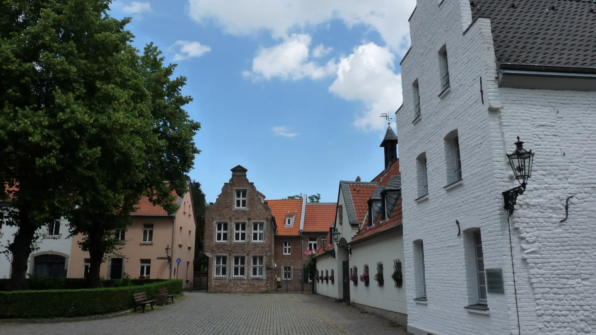Gepflasterter Innenhof mit alten Wohnhäusern und Bänken, umgeben von Bäumen.