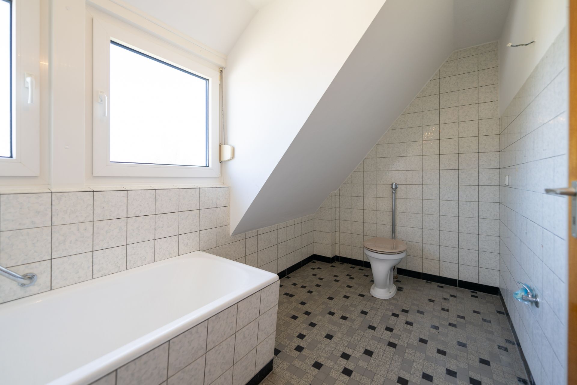 Helles Badezimmer mit Badewanne, Toilette und zwei Fenstern unter einer Dachschräge.