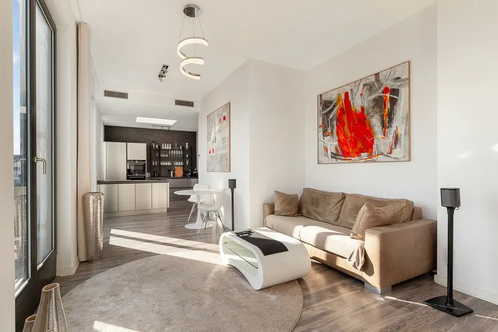 Modernes Wohnzimmer mit braunem Sofa, rundem Teppich, Kunst an der Wand und offener Küche im Hintergrund.