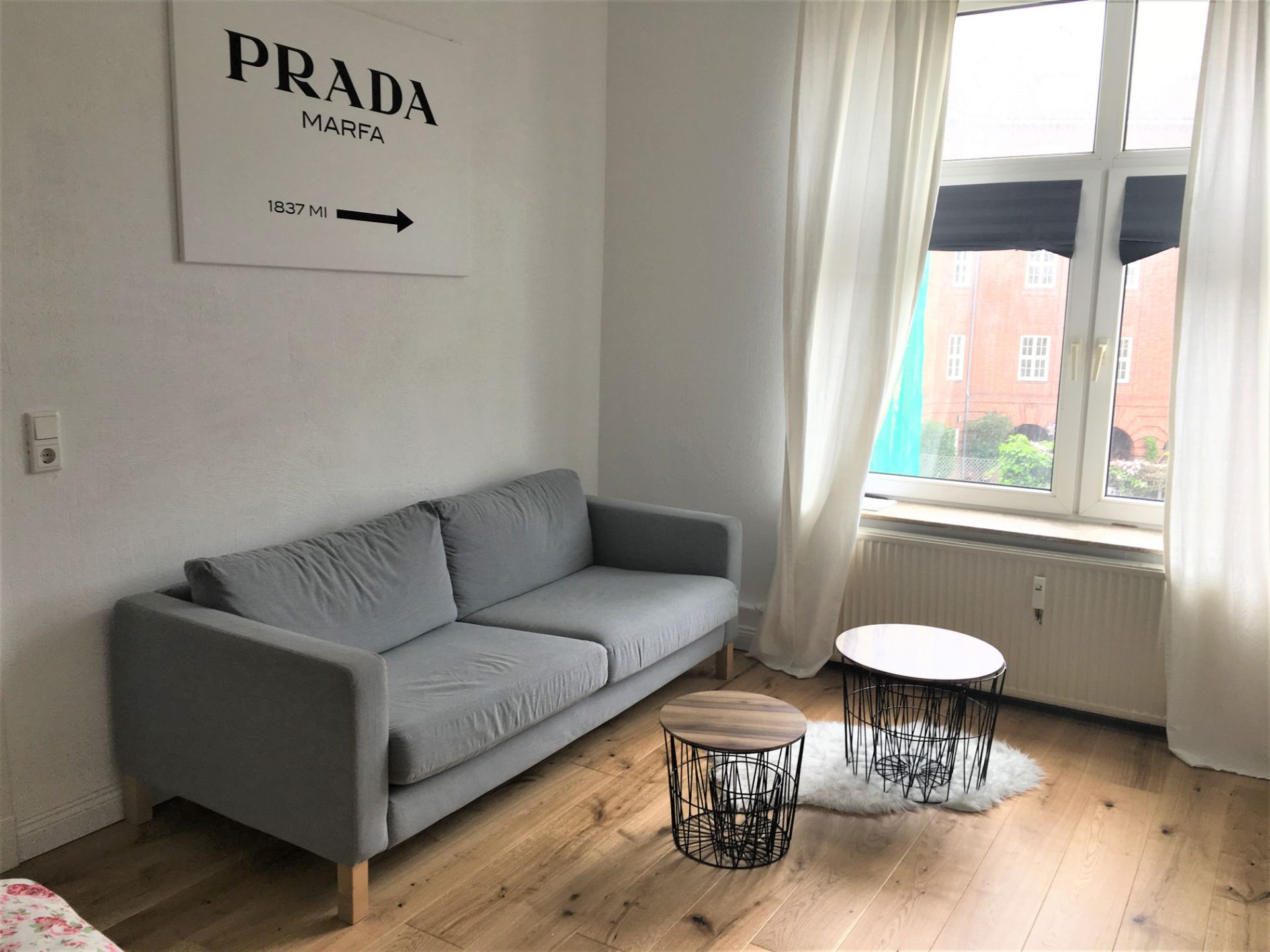 Helles Wohnzimmer mit grauem Sofa, zwei runden Tischen und großem Fenster mit weißen Vorhängen.
