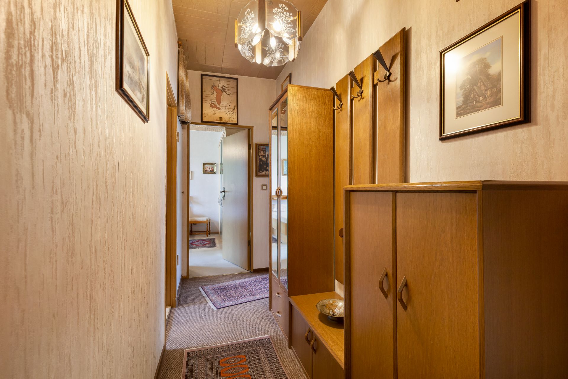Schmaler Flur mit Holzmöbeln, Teppichen, Bildern an den Wänden und einem Kronleuchter an der Decke.