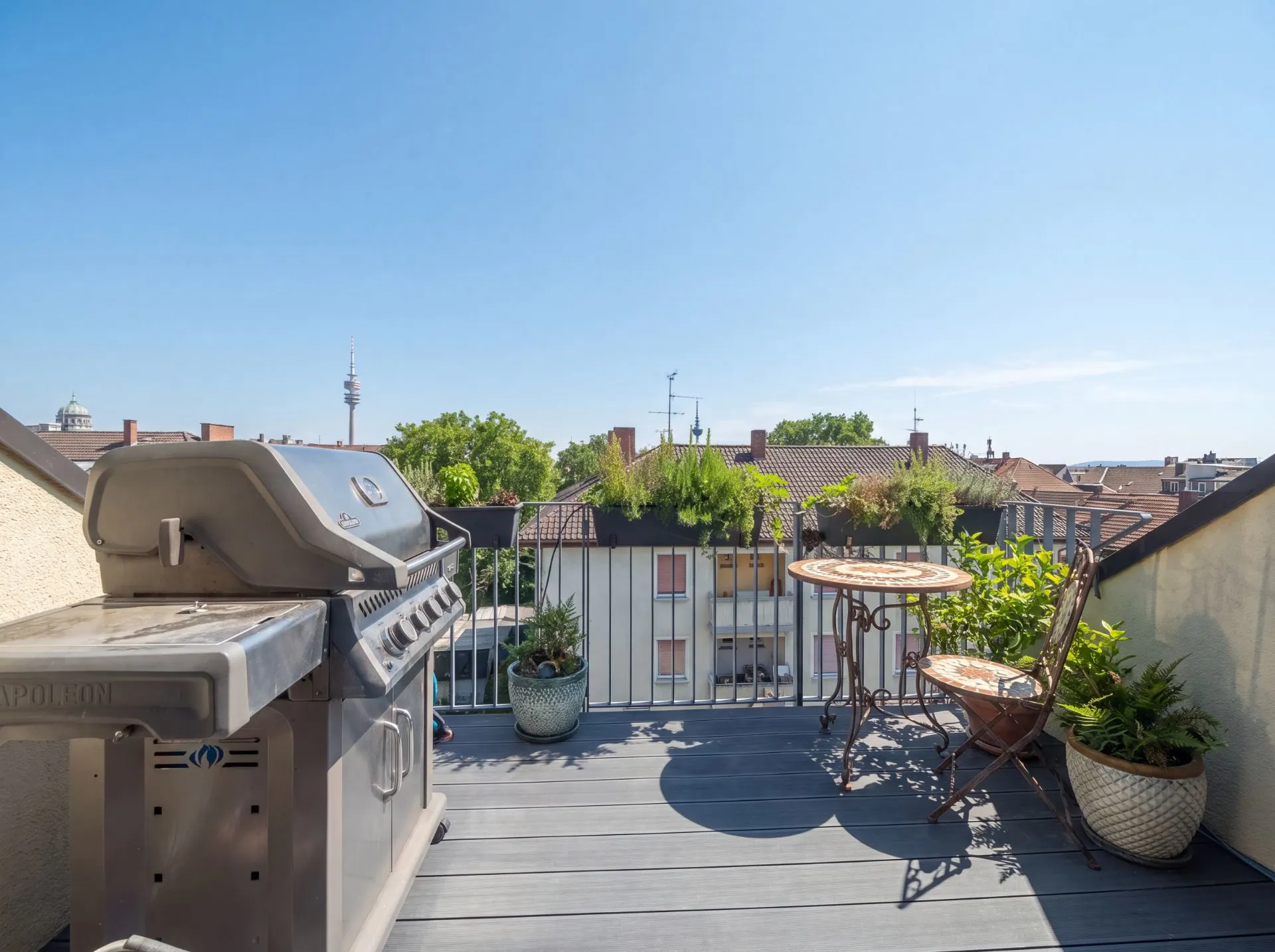 Balkon mit Grill, kleinem Tisch, zwei Stühlen und einigen Pflanzen, Blick auf Nachbarhäuser.