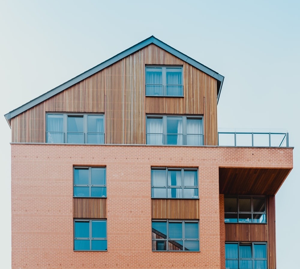 Modernes Wohngebäude mit roten Ziegeln und Holzverkleidung, viele Fenster, Balkon rechts.