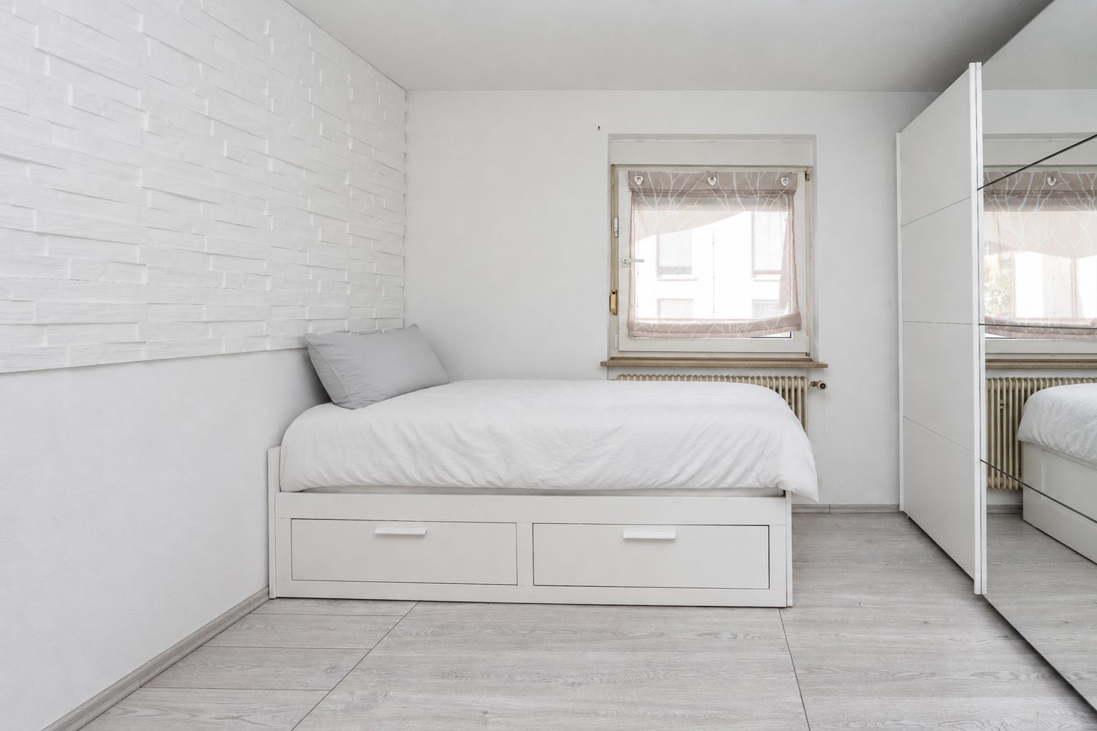Helles Schlafzimmer mit Einzelbett, Kissen, Fenster und weißem Kleiderschrank mit Spiegel.