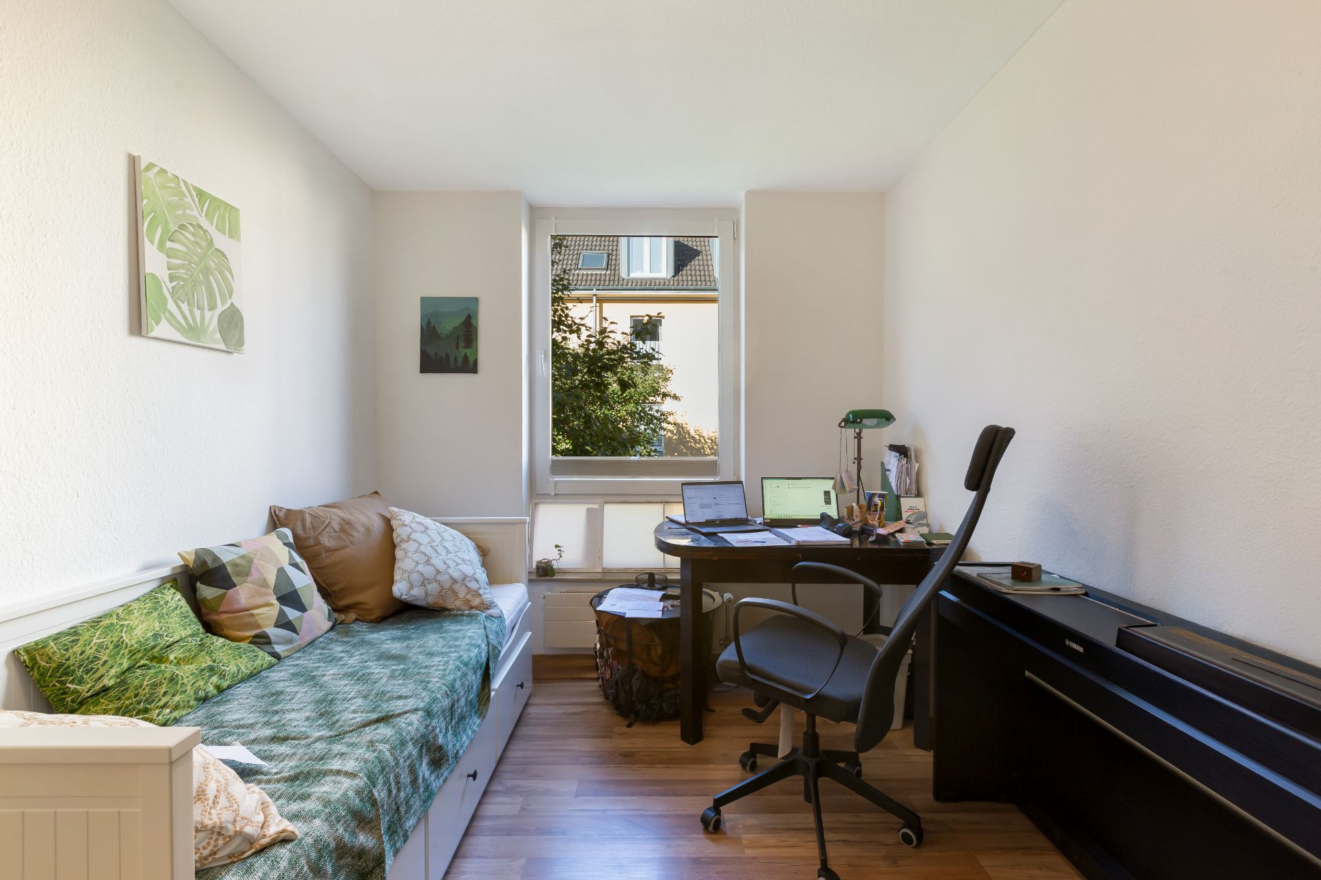 Helles Zimmer mit Einzelbett, Schreibtisch, Bürostuhl und Fenster mit Blick nach draußen.
