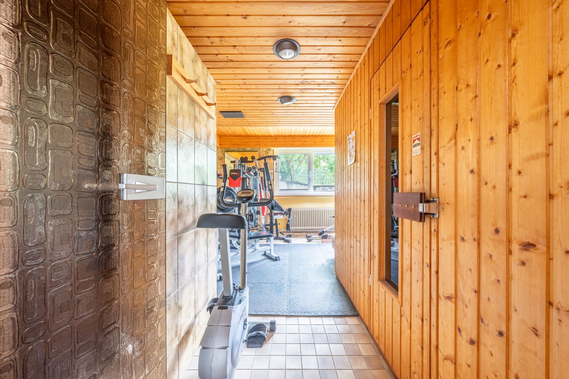 Flur mit Holzwänden, Sauna rechts und Fitnessgeräten im Hintergrund vor einem Fenster.