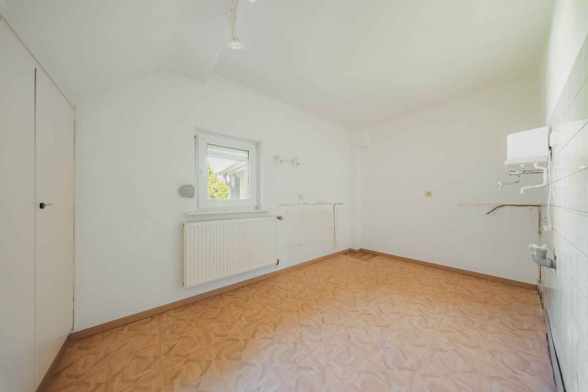 Leerer Raum mit Parkettboden, weißer Wand, kleinem Fenster und Heizkörper.