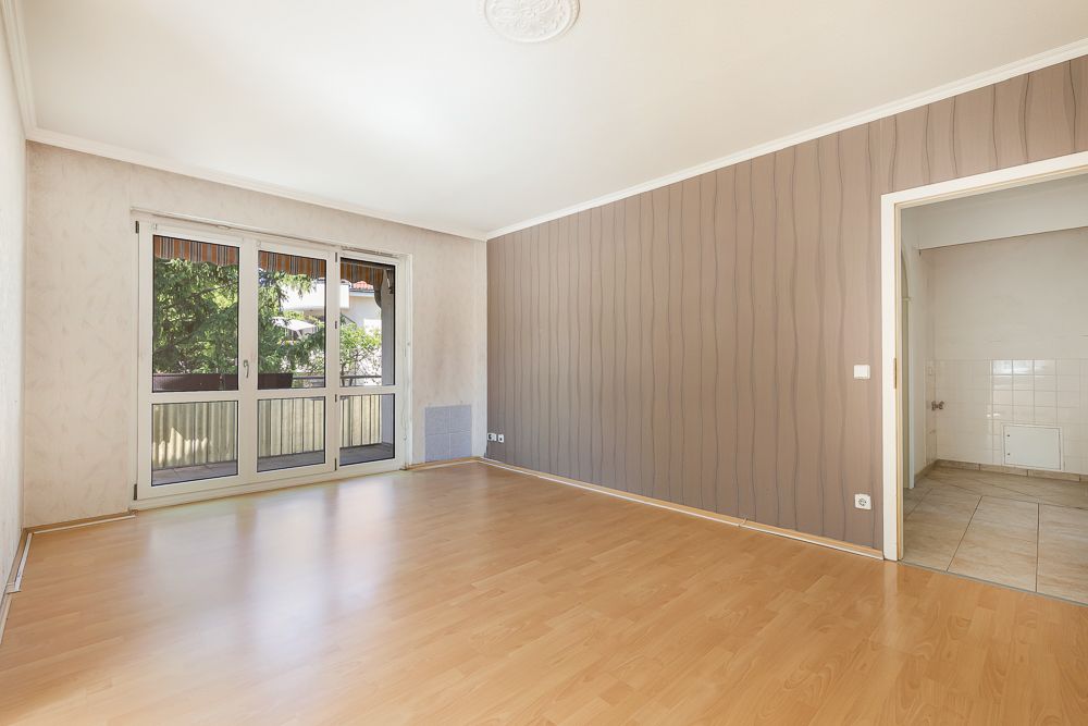 Helles Wohnzimmer mit Parkettboden, großem Fenster und Zugang zu einem angrenzenden Raum.