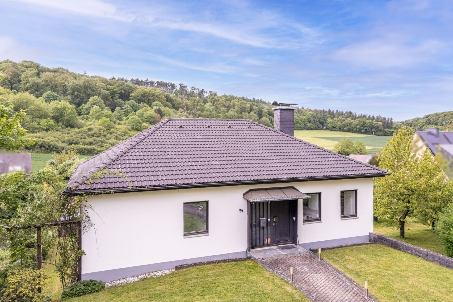 Einfamilienhaus mit dunklem Dach und weißer Fassade, umgeben von grüner Landschaft.