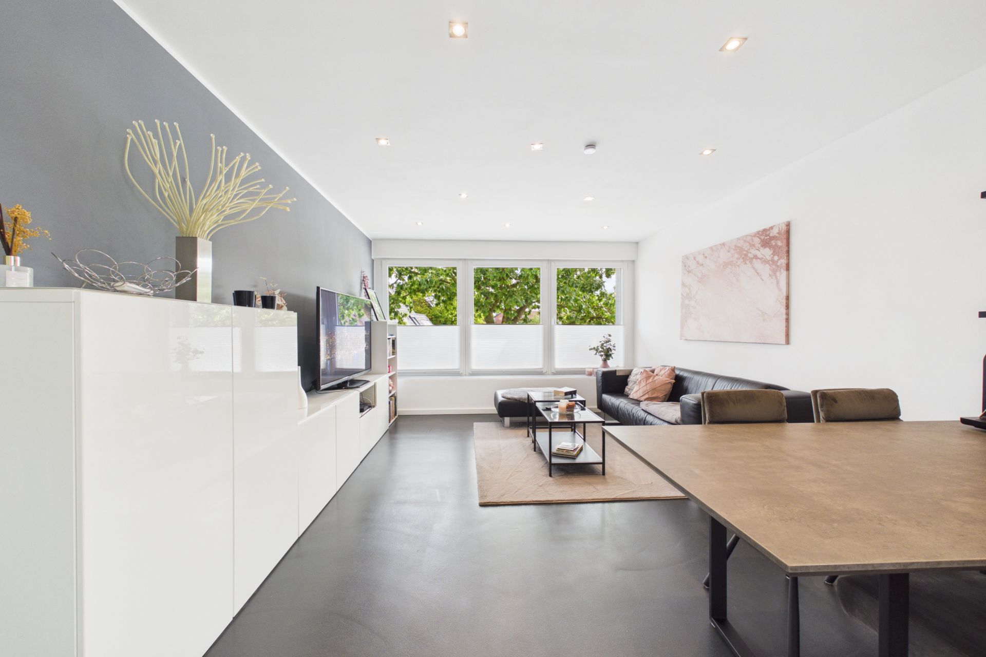 Modernes Wohnzimmer mit grauem Sofa, Esstisch, TV und großen Fenstern mit Blick ins Grüne.