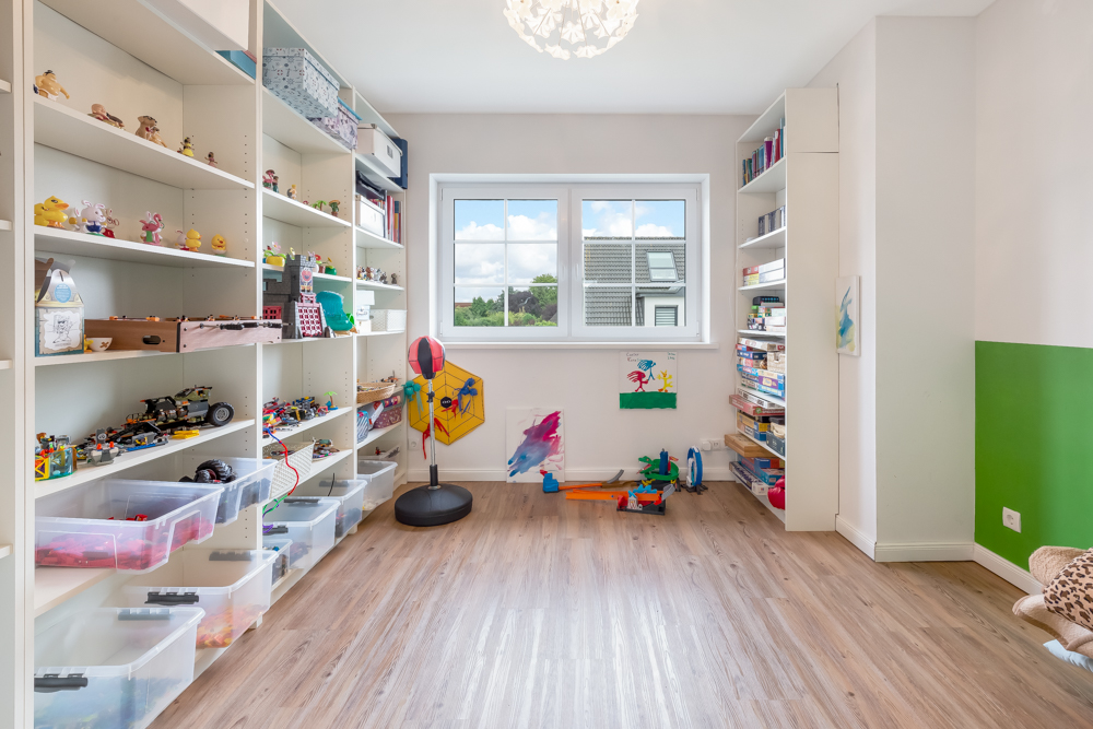 Helles Kinderzimmer mit Regalen voller Spielsachen und einem Fenster mit Blick nach draußen.