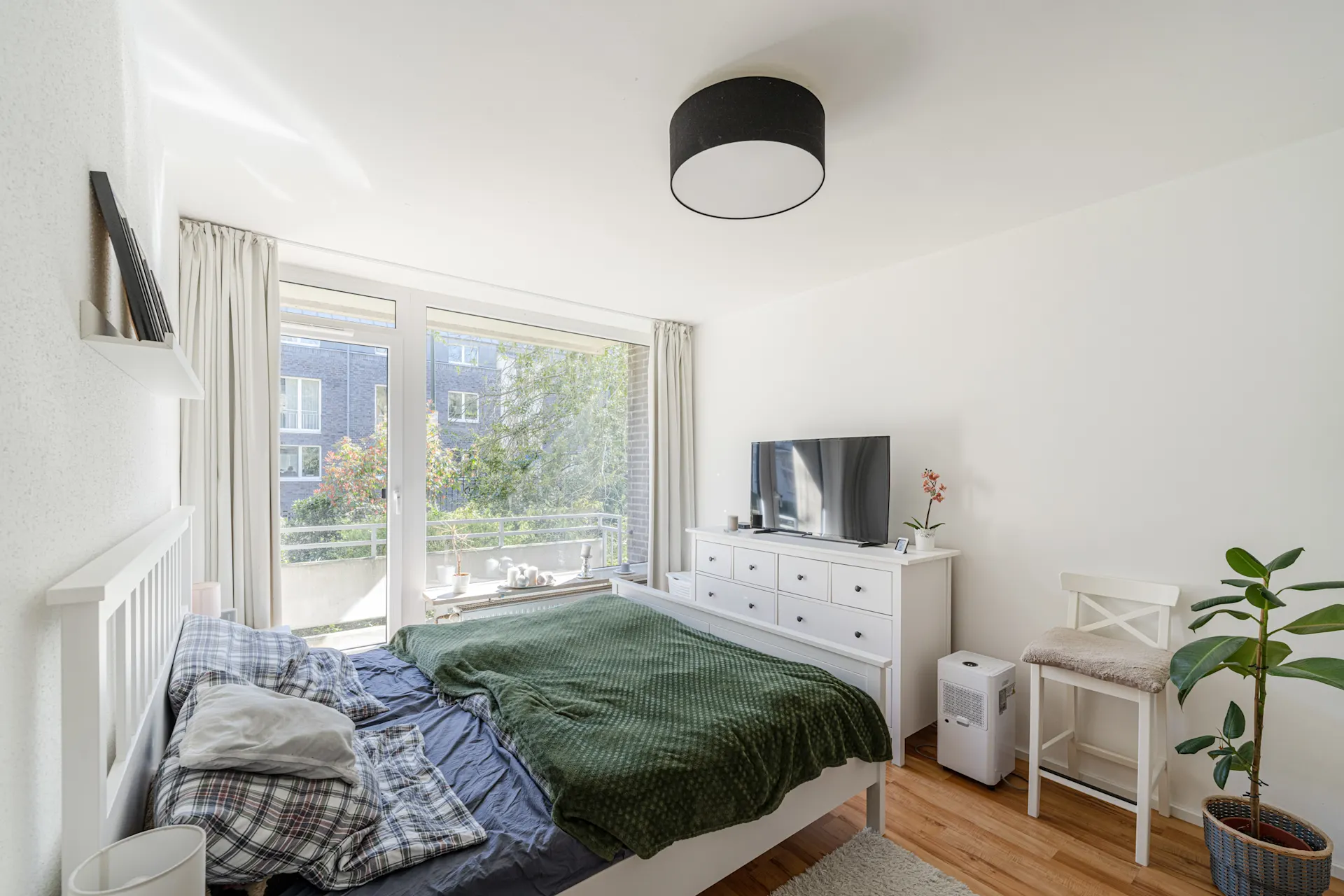 Helles Schlafzimmer mit Doppelbett, Kommode, Fernseher, Stuhl und großer Fensterfront.