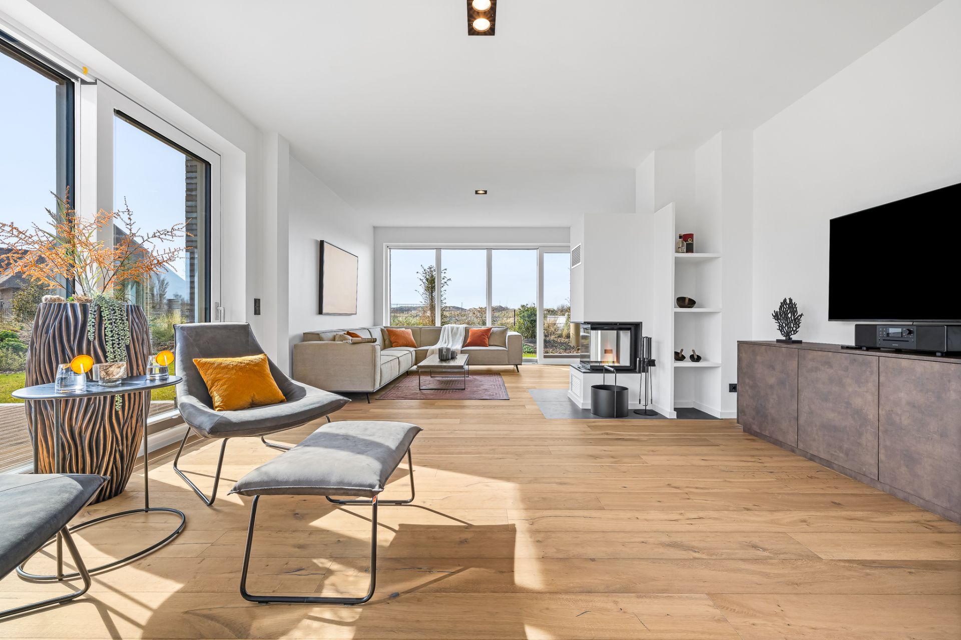 Modernes Wohnzimmer mit großen Fenstern, Kamin, Fernseher und gemütlichen Sitzmöbeln.
