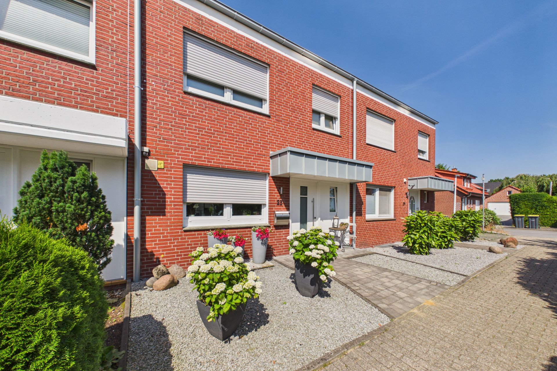 Reihenhaus mit roter Klinkerfassade, Rolläden und bepflanzten Blumenkübeln vor dem Eingang.