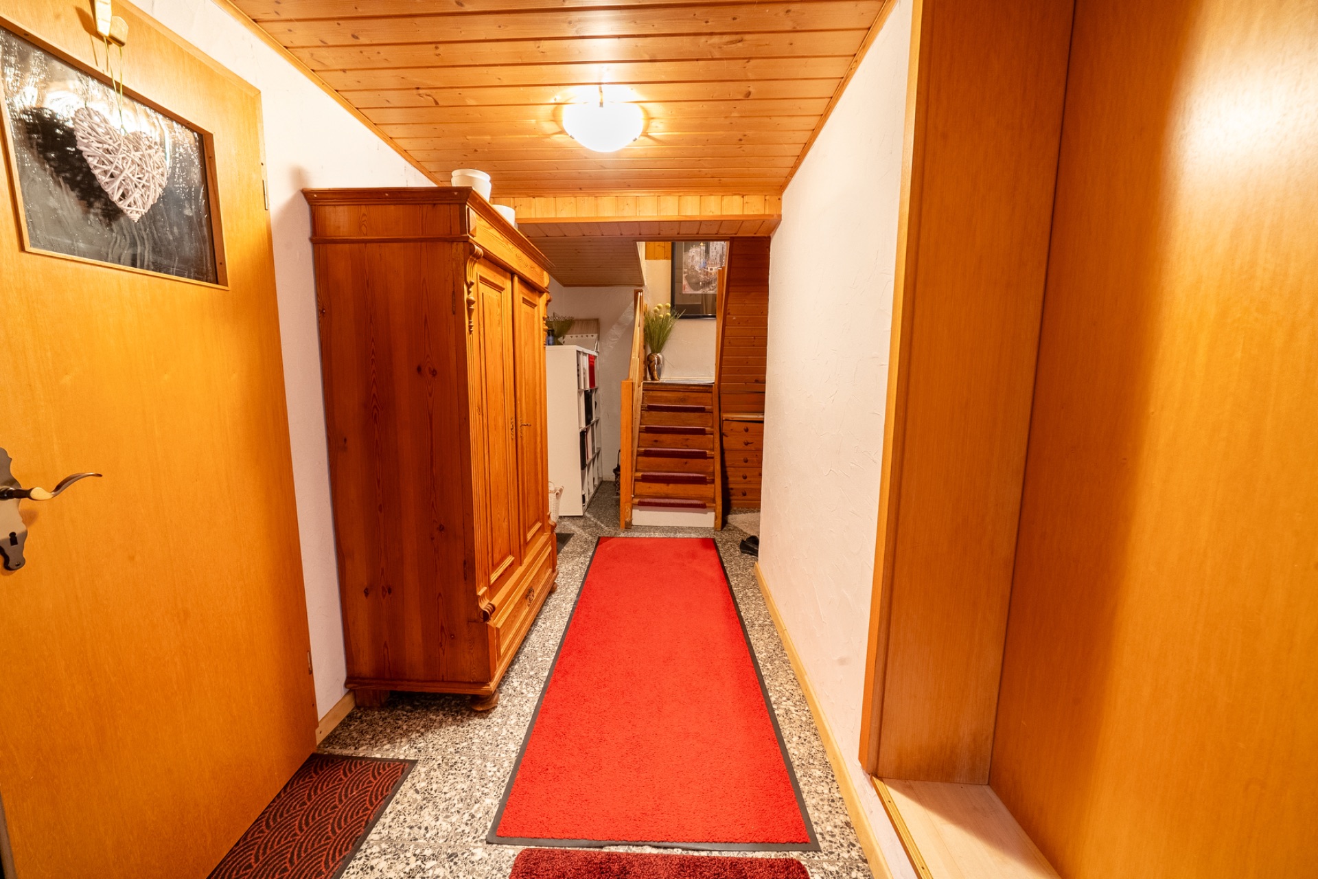 Heller Flur mit rotem Teppich, Holzschrank und Treppe im Hintergrund.