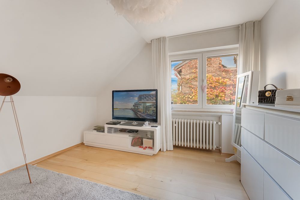 Helles Wohnzimmer mit Fernseher, Kommode, Fenster und Heizkörper auf hellem Holzboden.