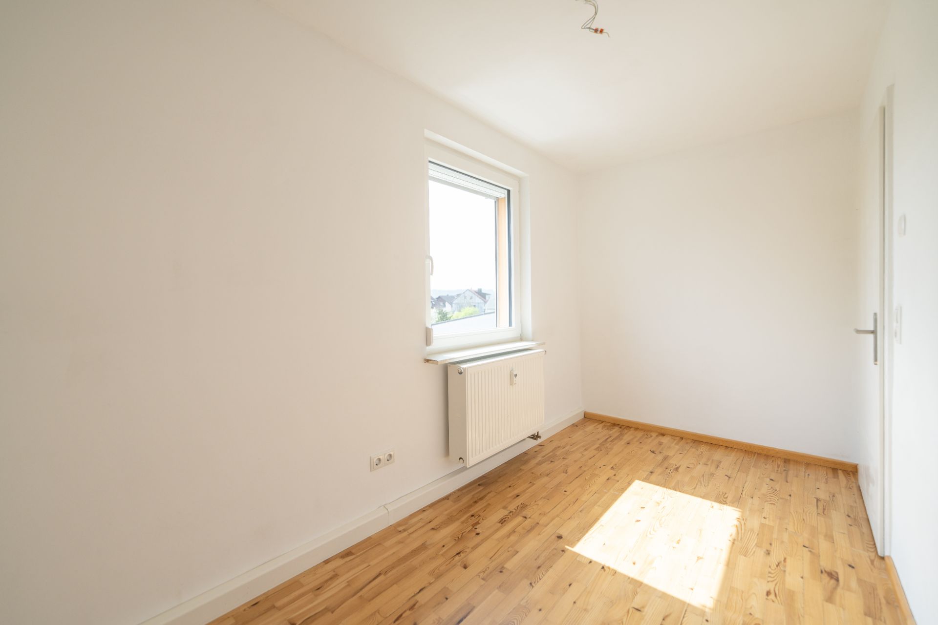 Helles, leeres Zimmer mit Holzfußboden, weißer Wand und einem Fenster mit Heizung darunter.