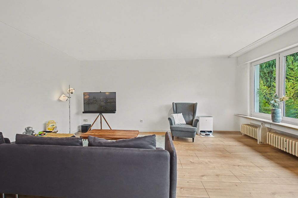 Helles Wohnzimmer mit grauem Sofa, Sessel, Couchtisch, Fernseher und großen Fenstern.