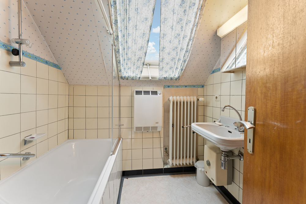 Kleines Badezimmer mit Badewanne, Waschbecken, Heizung und Dachfenster mit Vorhängen.