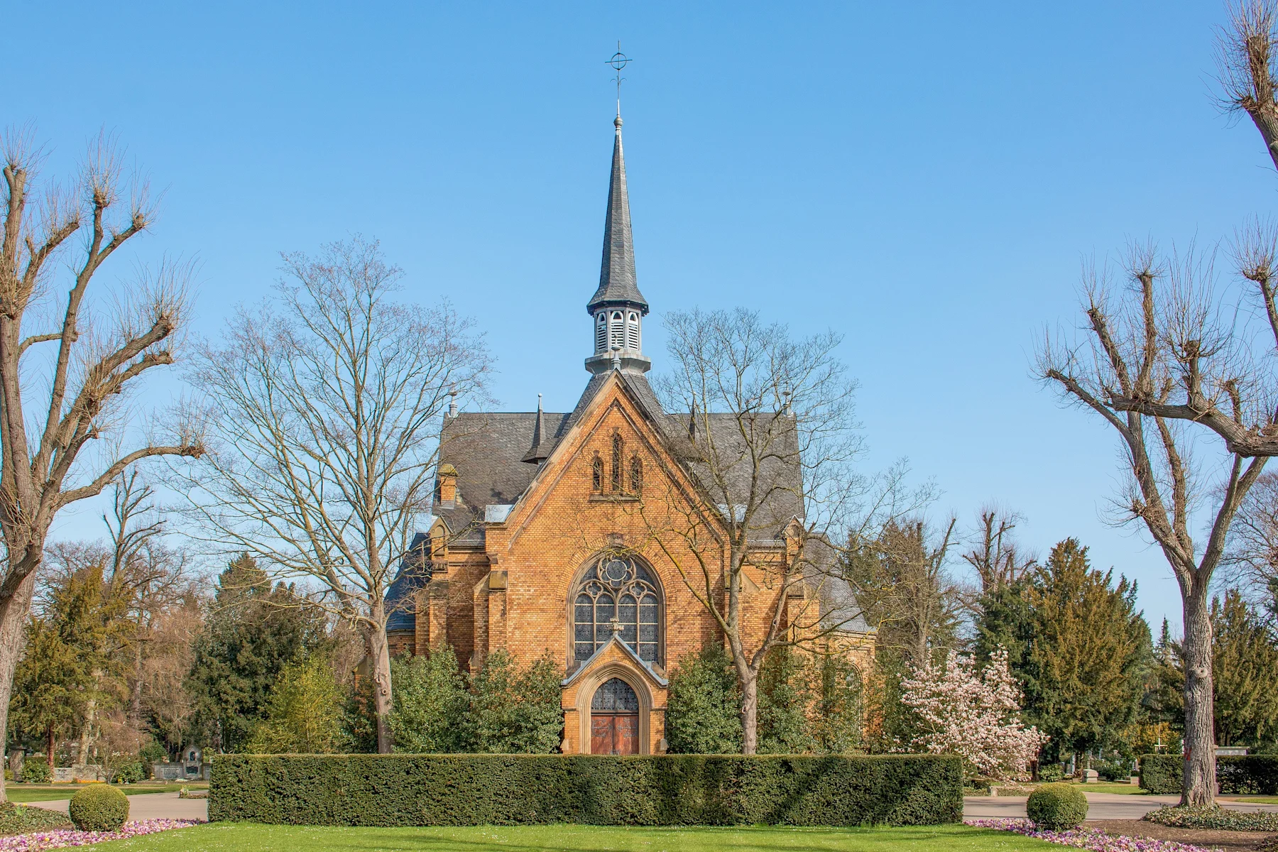 Backsteingebäude mit hohem Turm und großen Fenstern, umgeben von Bäumen und Hecken.