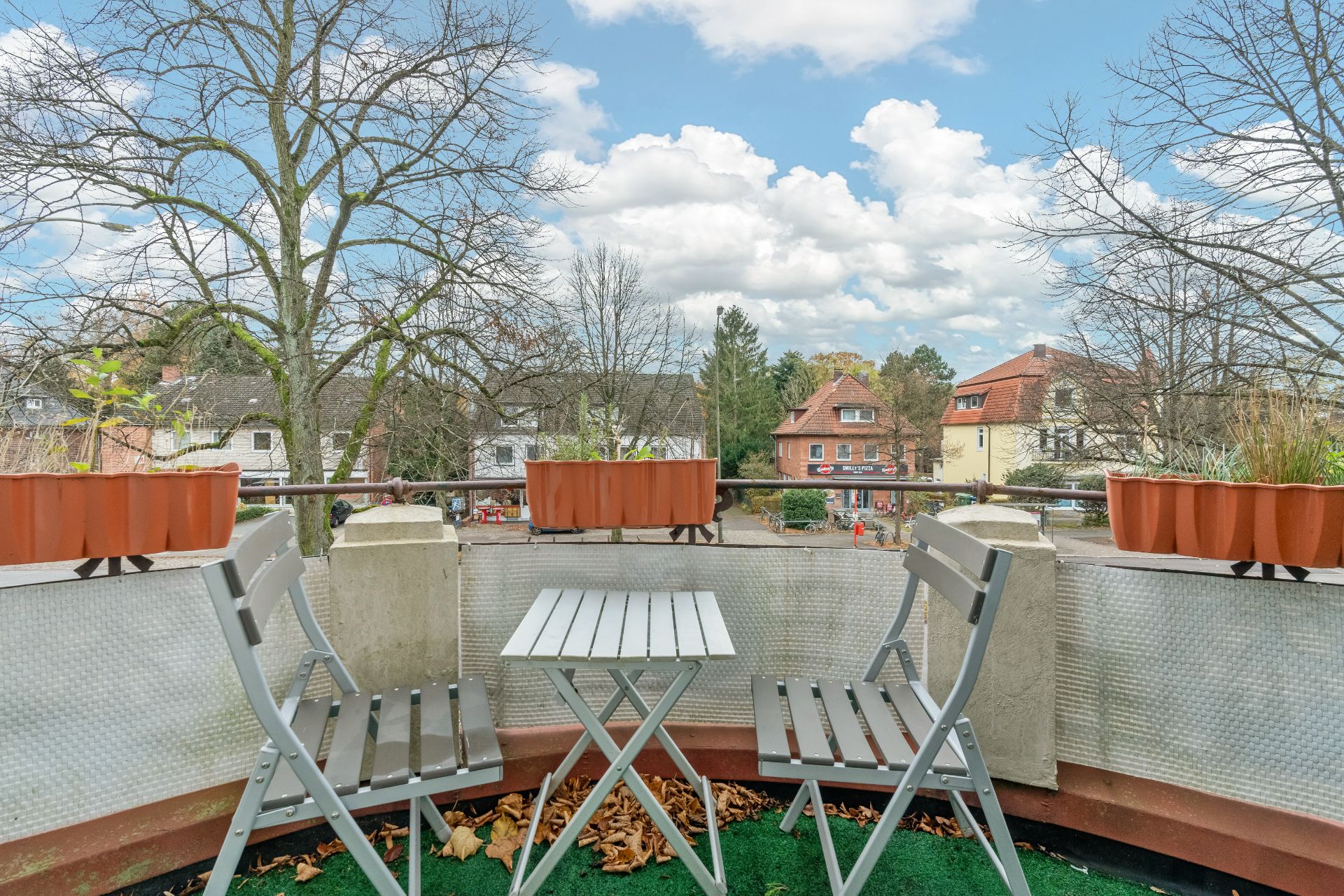 Balkon mit zwei Stühlen, Tisch und Blumenkästen, Blick auf Bäume und Wohnhäuser.