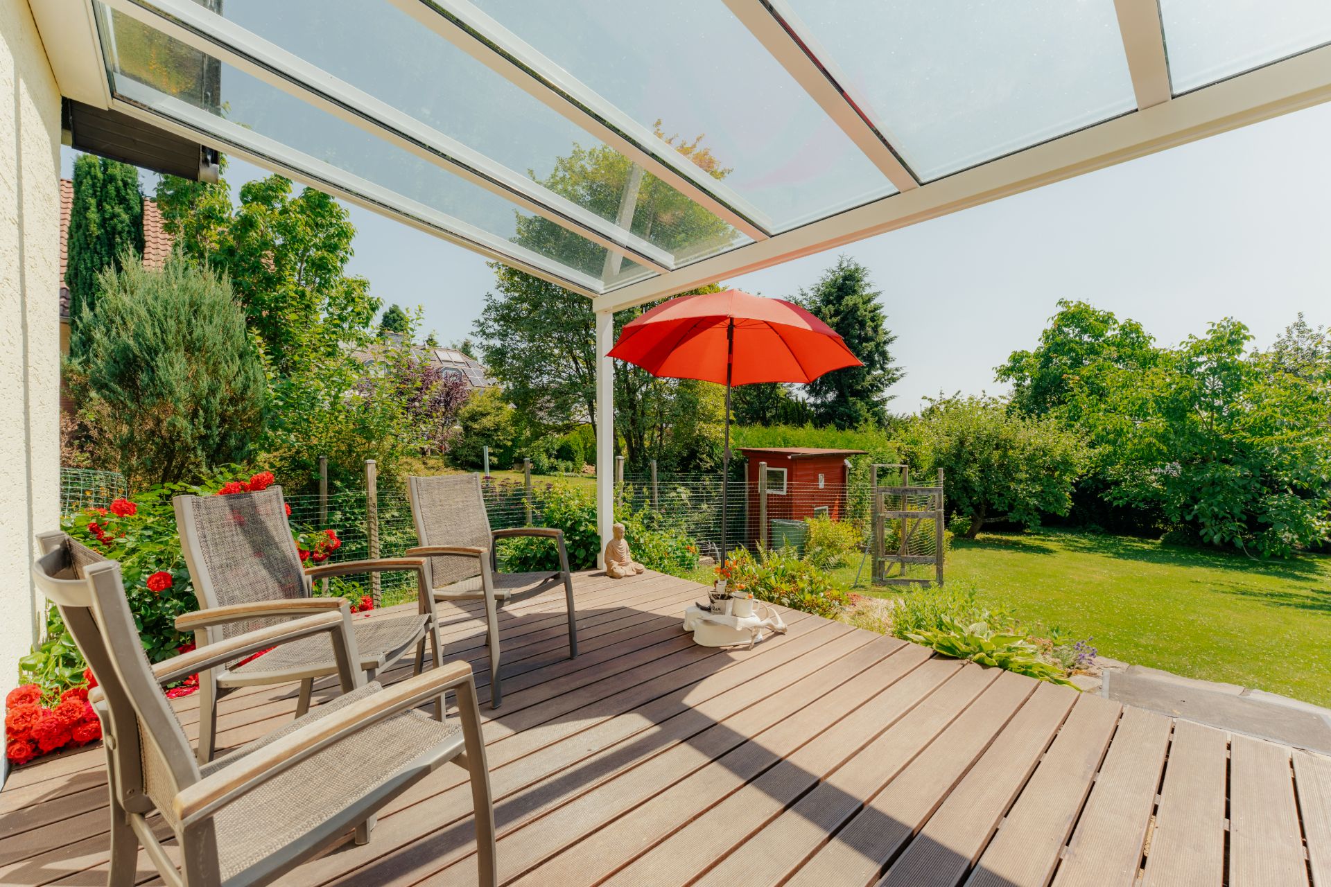 Überdachte Terrasse mit drei Stühlen, rotem Sonnenschirm und Blick in den Garten.