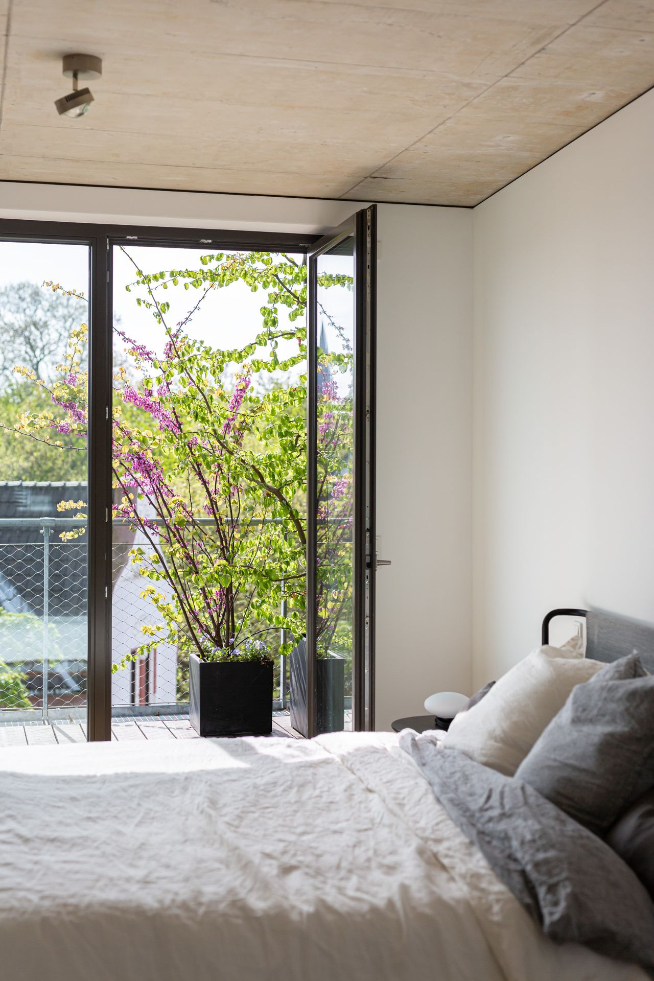 Helles Schlafzimmer mit Bett, offenen Glastüren und einer Pflanze auf dem Balkon.