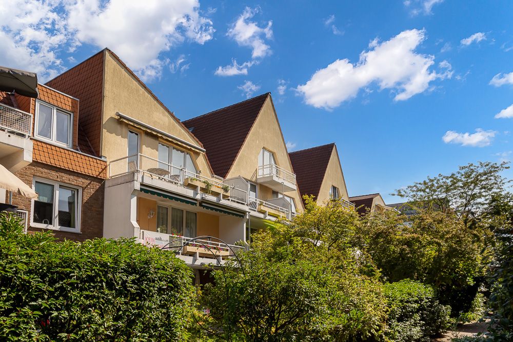 Mehrfamilienhaus mit Balkonen und Garten, spitz zulaufende Dächer, blauer Himmel mit Wolken.