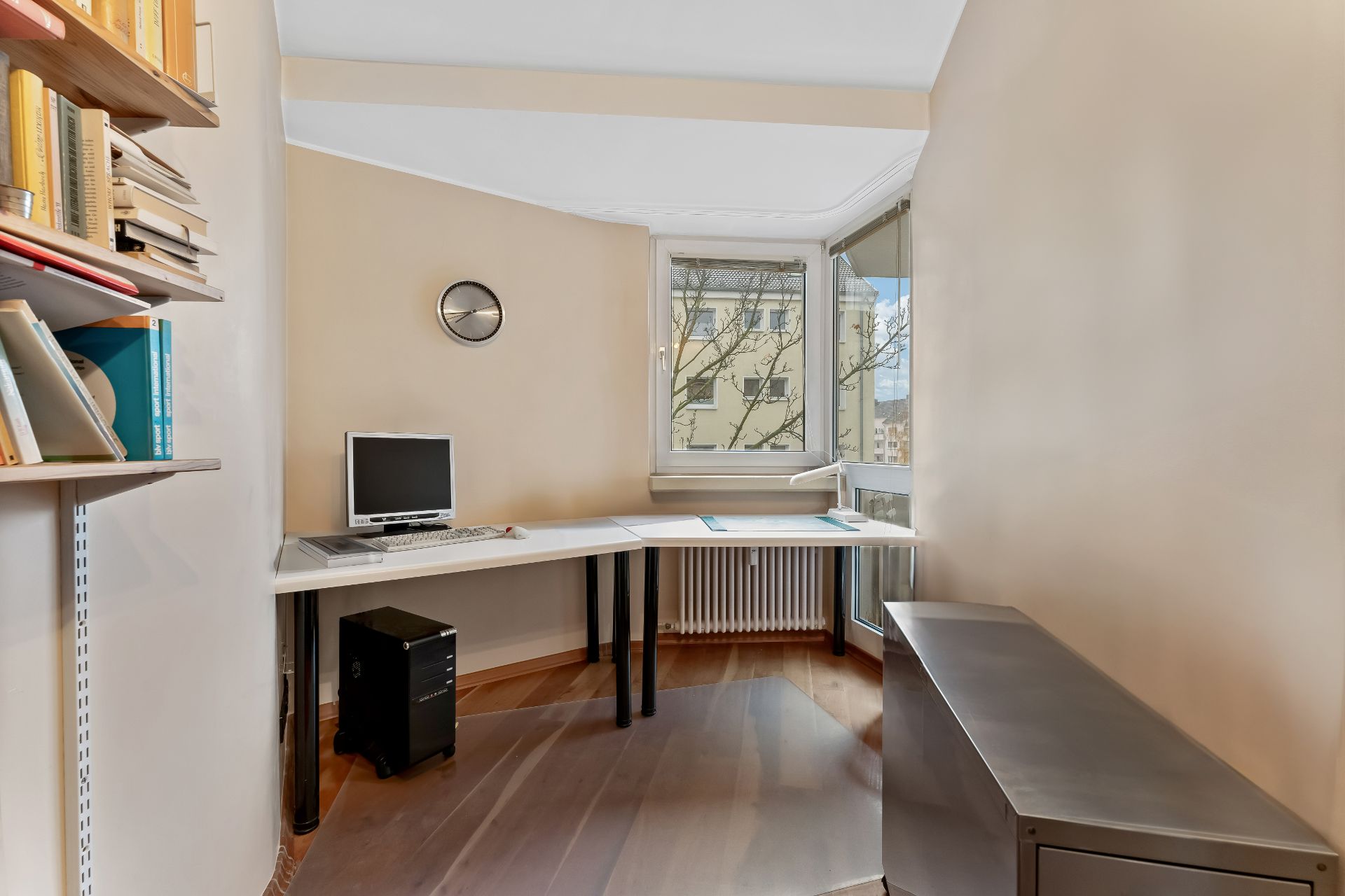 Helles Arbeitszimmer mit großem Schreibtisch, Computer, Bücherregal und Fenster mit Blick nach draußen.