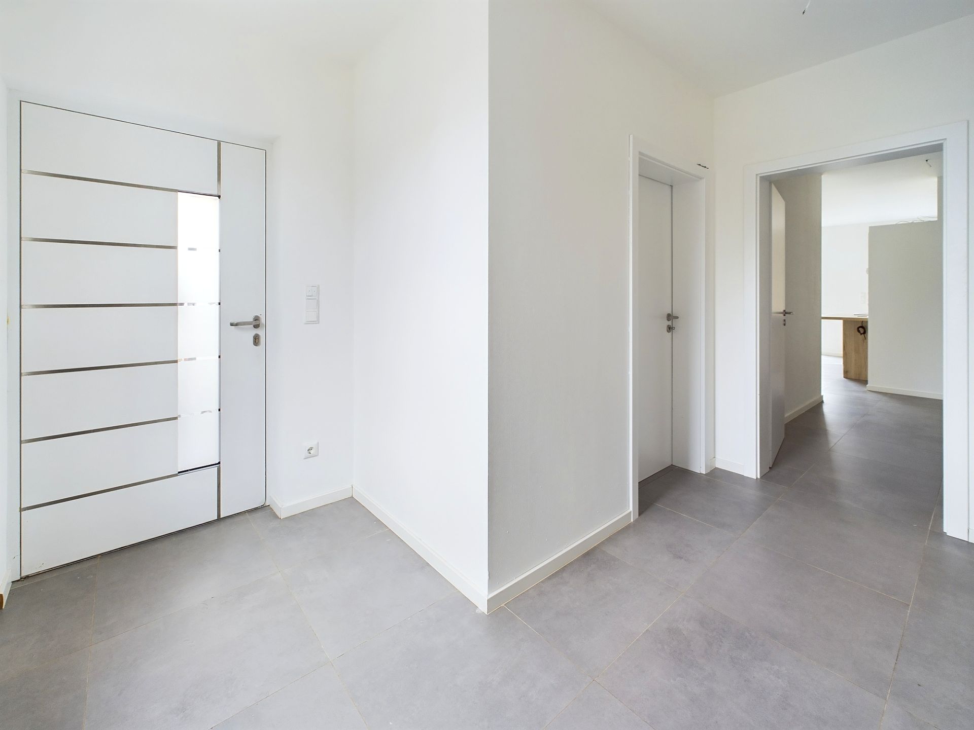 Heller Flur mit grauen Bodenfliesen und weißer Eingangstür in einem Wohnhaus.