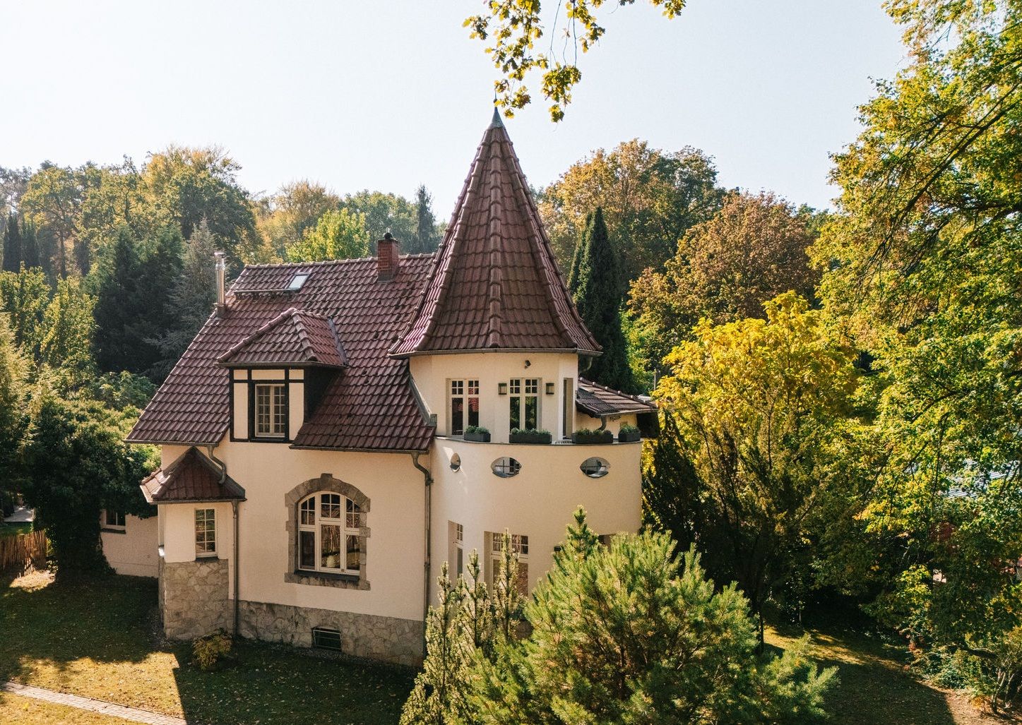 Helles Wohnhaus mit spitzem Turmdach und roten Ziegeln, umgeben von Bäumen und Garten.