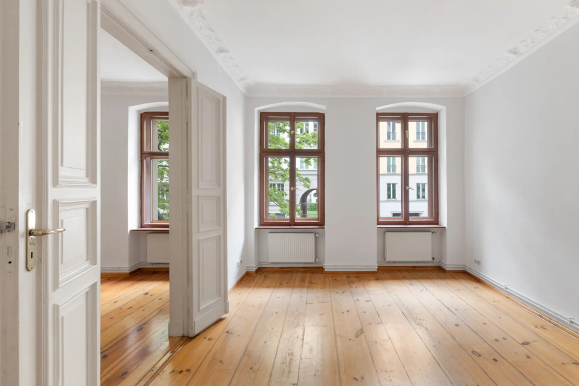 Helles leeres Zimmer mit Holzboden, weißen Wänden und drei großen Fenstern.