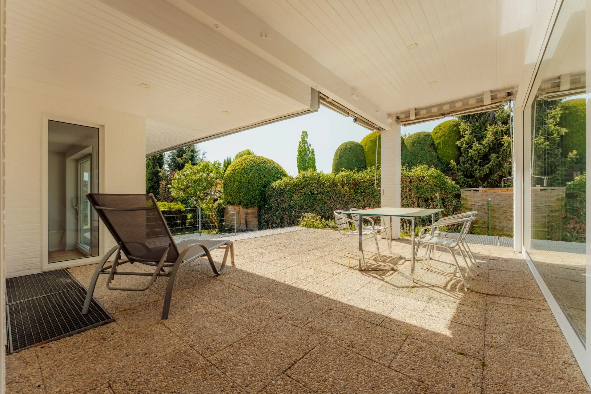 Überdachte Terrasse mit Liegestuhl, Tisch und Stühlen, Blick auf Garten mit Sträuchern.