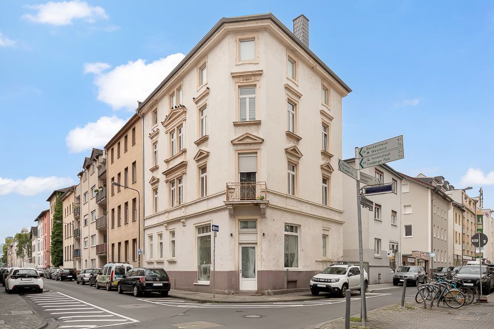 Eckhaus mit drei Stockwerken an einer Straßenecke, mehrere geparkte Autos und Fahrräder sichtbar.
