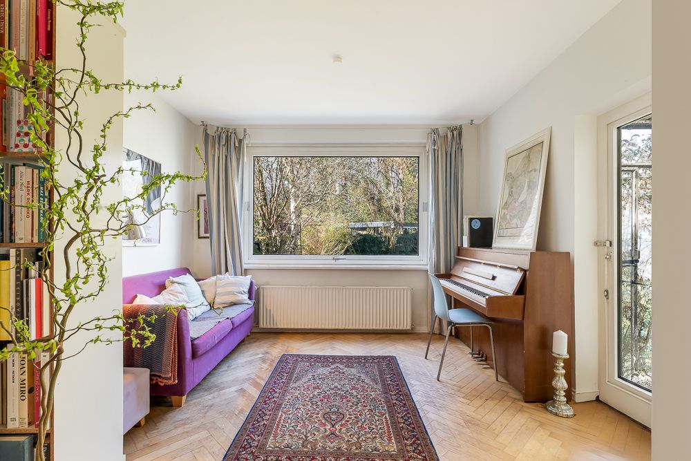 Wohnzimmer mit lila Sofa, Teppich, Klavier, Bücherregal und großem Fenster mit Blick ins Grüne.