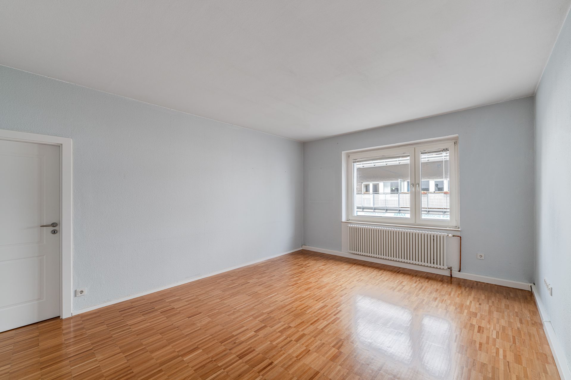 Leeres Zimmer mit Holzboden, weißer Tür und Fenster mit Heizkörper darunter.